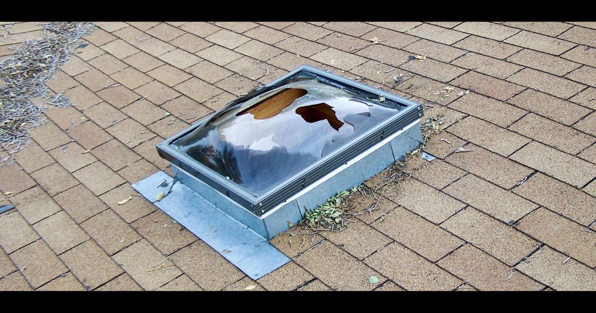 A residential roof skylight with a punctured and shattered dome cover — visible storm impact damage that compromises the seal and exposes the home interior to water intrusion